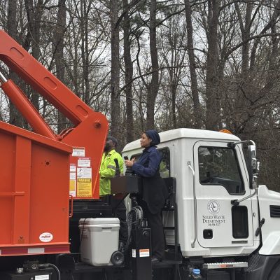 Workers loading trash into a garbage truck in a wooded area.
